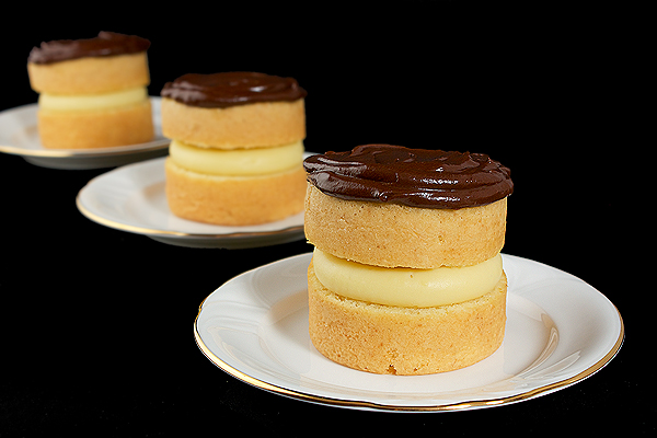 Boston Cream A photo of three Boston Cream desserts displayed in a row on white dessert dishes with a black background.
