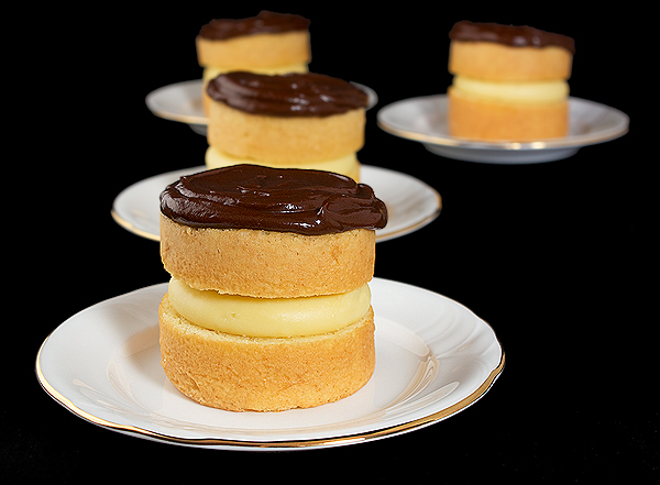 chocolate ganache, chocolate glaze, boston cream recipe A photo of four Mini Boston Cream Pie cakes served on white dessert dishes with a black background.