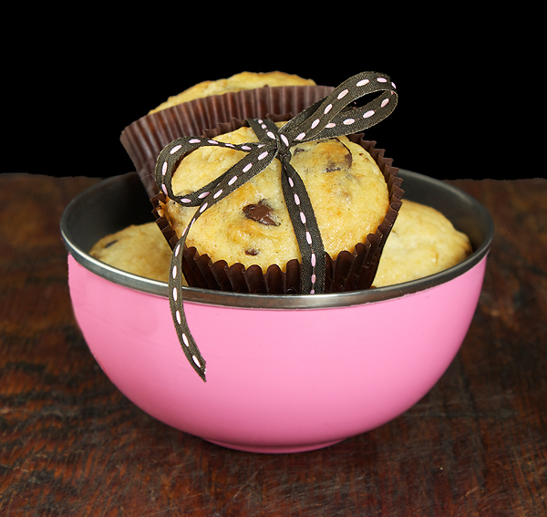Banana Muffins with Chocolate Chips A photo of 4 small Banana Muffins with Chocolate Chips in a small pink bowl and one of the muffins has a ribbon brown and pink ribbon tied around it.