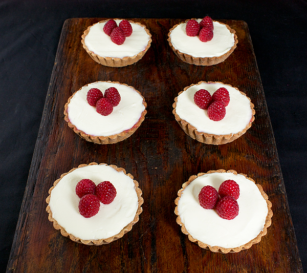 Hazelnut, Raspberry Jam and White Chocolate Ganache Tarts A photo taken looking down on six Hazelnut, Raspberry Jam and White Chocolate Ganache Tarts garnished with fresh raspberries.