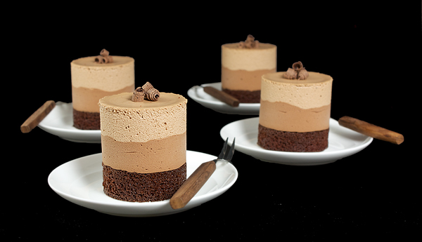 Chocolate Mousse Cakes A photo of 4 Chocolate Mousse Cakes layered with Dark Chocolate Cake, Bittersweet Chocolate Mousse and Milk Chocolate Mousse displayed on small white dessert dishes with dessert forks with a black background.