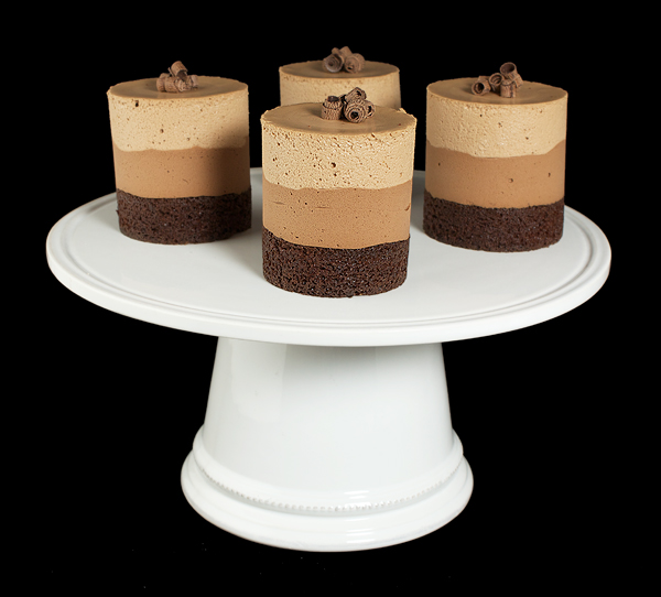 Mousse Cakes A photo of 4 Mousse Cakes garnished with Dark Chocolate Curls displayed on a white cake stand.