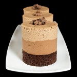 A photo of 3 Triple Chocolate Mousse Cakes displayed in a row on a white rectangular dish with a black background.