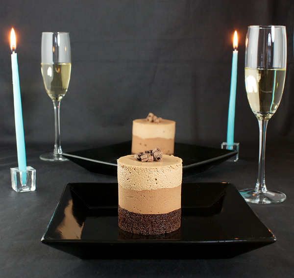 Chocolate Mousse Cakes A photo of dessert for two, Chocolate Mousse Cakes garnished with chocolate curls on black dessert plates with 2 champagne filled glass flutes with baby blue lit candles with a black background.