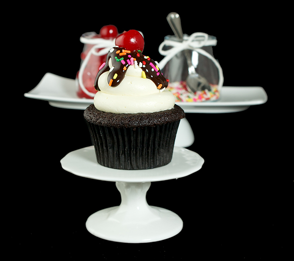 cupcake decorating ideas, best chocolate cupcakes, vanilla frosting recipe A photo a Hot Fudge Sundae Cupcake on a white cupcake stand with a jar of maraschino cherries and rainbow nonpareils in behind on a white rectangular dish.