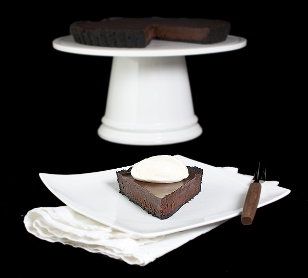 Oreo Chocolate Truffle Tart with Chocolate Glaze A photo of an Oreo Chocolate Truffle Tart with Chocolate Glaze displayed in the background on a white cake stand with a slice of Oreo Chocolate Truffle Tart on a white dessert dish placed in front of the cake stand.