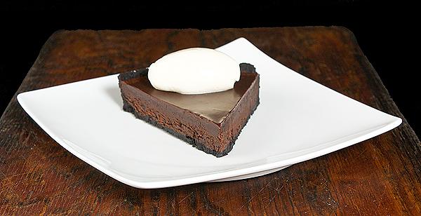 Chocolate Tart A photo of a slice of Chocolate Tart with a quenelle of Mascarpone Cream on a white dish placed on a dark wooden board.
