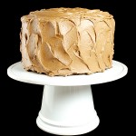 Chocolate Cake with Caramel-Milk Chocolate Frosting A photo of a Chocolate Cake with Caramel-Milk Chocolate Frosting displayed on a white cake stand with a black background.