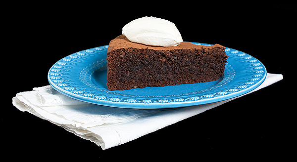 A photo of a slice of Torta Caprese with a quenelle of Sweetened Whipped Cream and a dusting of cocoa placed on a blue dessert dish.