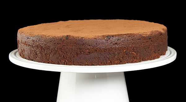 A photo of a Torta Caprese, Italian Flourless Chocolate Torte dusted with cocoa powder and displayed on a white cake stand.