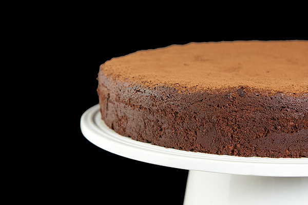 A cropped photo of an Italian Torta Caprese dusted with cocoa and displayed on a white cake stand.