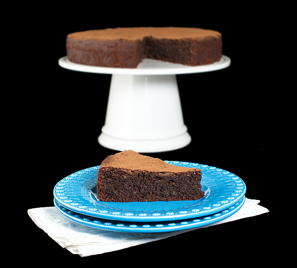 A photo of Italian Cake, Torta Caprese displayed on a white cake stand with a slice removed and displayed on a blue dessert dish set in front.