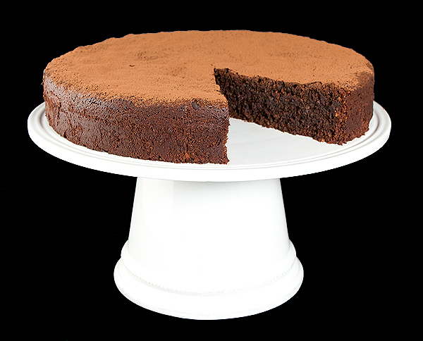 A photo of an Italian Flourless Chocolate Cake, Torta Caprese with a slice removed displayed on a white cake stand.