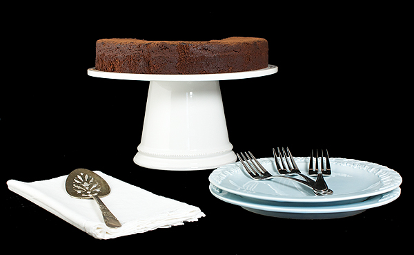 A photo of a Torta Caprese displayed on a white cake stand along side pale blue dessert dishes, dessert forks and a vintage cake lifter on a white cloth napkin.