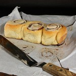A photo of 3 Cinnamon Rolls fresh out of the oven on non-stick baking paper with an offset spatula off to the side.