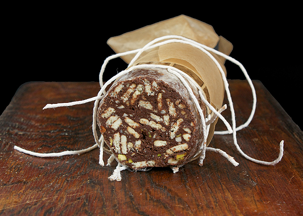 salame chocolate, salami di chocolate, chocolate dessert recipes A photo of a Salame al Cioccolato (Chocolate Salami) wrapped in non-stick baking paper secured with string on a dark wooden cutting board.