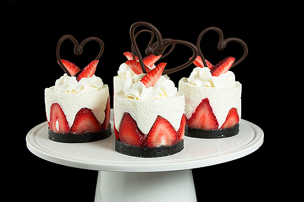 A photo of an Italian Cake, Torta di Fragole e Crema Soffice di Yogurt, in mini form, garnished with fresh strawberries and a chocolate heart displayed on a white cake stand.