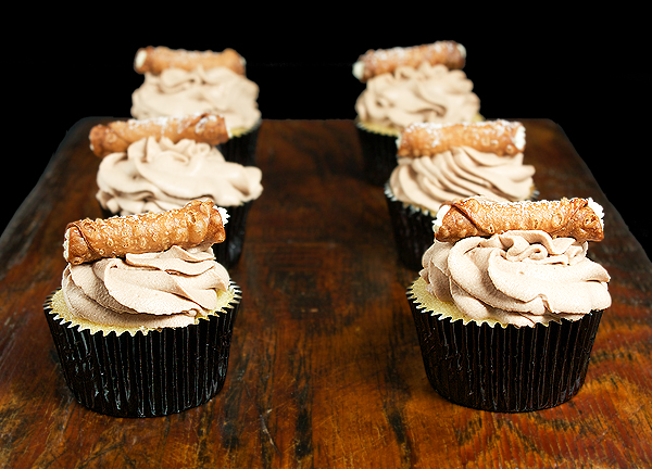 Vanilla Cupcakes A photo of 6 Cannoli Cupcakes decorated with mini cannoli displayed on a dark wooden board.