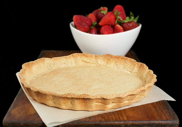 A photo of a baked Pasta Frolla tart shell and a bowl of fresh strawberries on a dark wooden board.