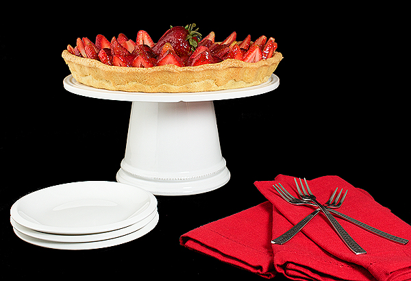 A photo of a Strawberry Tart with Pastry Cream lightened with Whipped Cream and topped with fresh Strawberries and coated with a thin layer of Apricot Glaze, displayed on a white cake stand with dessert dishes, dessert forks and red napkins.