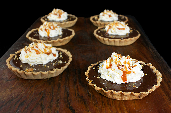 Chocolate-Caramel Tart {Tartellette al Cioccolato e Caramello} A photo of six Chocolate-Caramel Tarts {Tartellette al Cioccolato e Caramello} displayed on a wooden board.