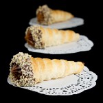 Italian Pastry, Cannoncini di Sfoglia A photo of three Cannoncini di Sfoglia con Crema Pasticcera al Cioccolato (Pastry Horns filled with Chocolate Pastry Cream) place on white doilies and displayed in a row, one behind the other.
