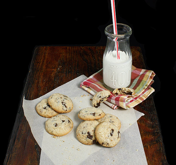 Grace's Sweet Life Hazelnut Cookies 1 A photo of Italian hazelnut cookies displayed on sheets of parchment placed on a dark wooden board and served with a glass of milk resting on a red check cloth napkin.