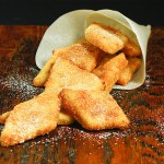 A photo of Crema Fritta alla Veneziana, Venetian Style Fried Cream, dusted with confectioners' sugar in a paper cone displayed on a dark wooden board.