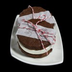 A photo of three Chocolate Sablée-Gelato Ice Cream Sandwiches wrapped in a strip of parchment paper with red and white string displayed on a white rectangular-shaped dessert dish.