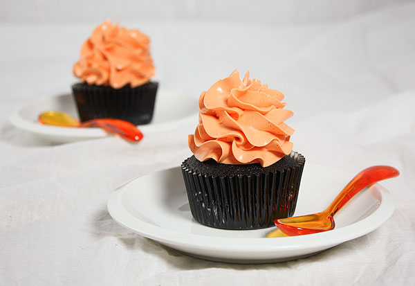 Grace's Sweet Life Pumpkin Chocolate Cake A photo of two mini Pumpkin Chocolate Cakes decorated with orange-tinted frosting on white dessert dishes displayed with orange spoons.