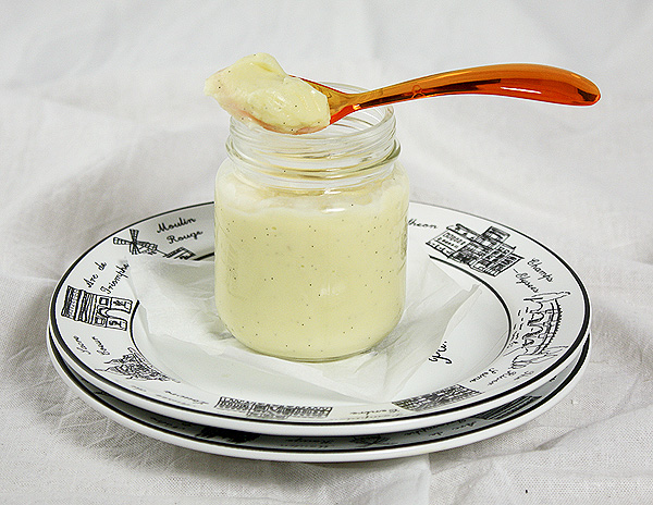 Close up photo of White Chocolate Pudding in a glass jar displayed on a stack of dessert dishes with an orange acrylic spoon.