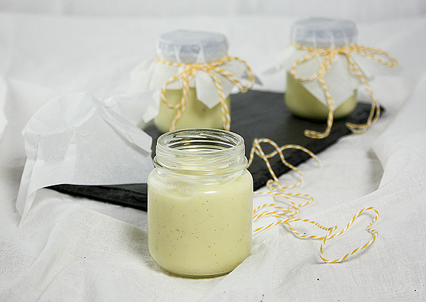 A photo of White Chocolate Vanilla Bean Pudding displayed in glass jars topped with non-stick baking paper squares secured with orange and white string.