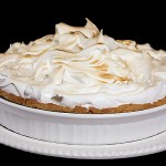 A photo of Lemon Meringue Tart in a white ceramic tart dish displayed on a white cake stand with a black background.
