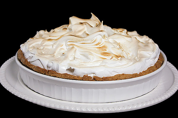 A photo of Lemon Meringue Tart in a white ceramic tart dish displayed on a white cake stand with a black background.