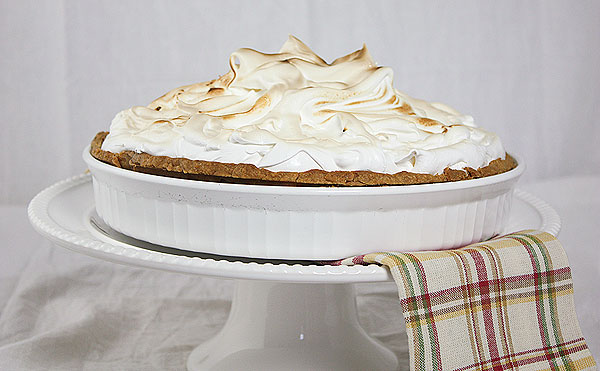 A photo taken of Lemon Meringue Tart in a white tart dish displayed with a multicoloured cloth napkin displayed on white cake stand.
