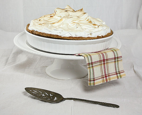 A photo of Lemon Meringue Tart in white ceramic tart dish displayed on a white cake stand with red striped napkin and antique metal cake server.