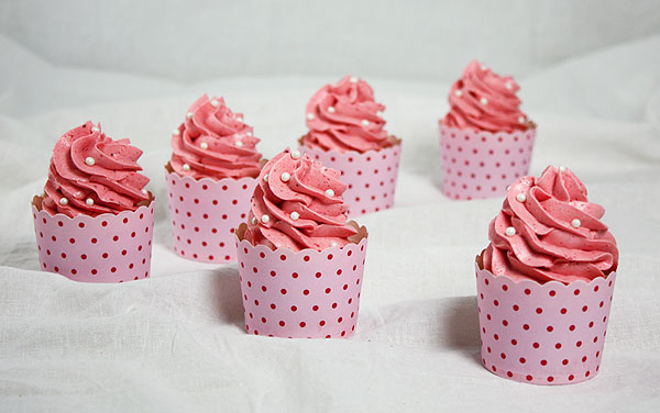 A photo of Gourmet Strawberry Cupcakes with Pink Strawberry Meringue Buttercream frosting decorated with white sugar pearlsin red dotted, pink cupcake cups placed in a v-formation on white cloth.