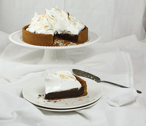 A photo of S mores Pie displayed on white cake stand with a slice of pie displayed in front on double stacked white dessert dishes.