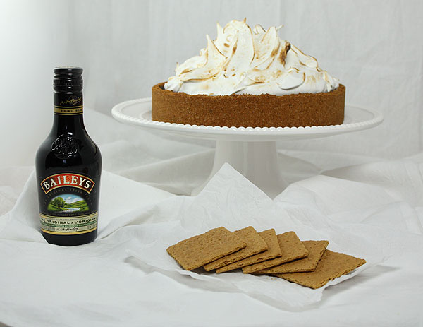 A photo of Spikes Smores Pie displayed on white cake stand behind bottle of Baileys and graham crackers.
