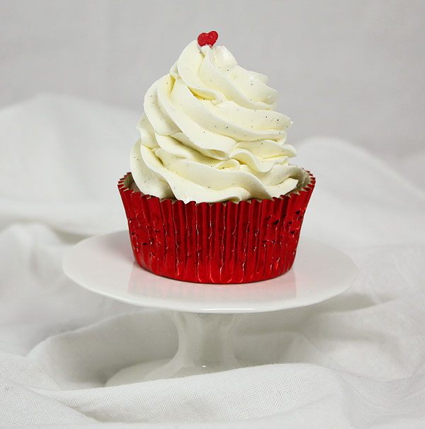 A photo of a Strawberry Cupcake with Swiss Meringue Buttercream in red foil cupcake liner decorated with a single red heart sprinkle on top displayed on white cake stand.
