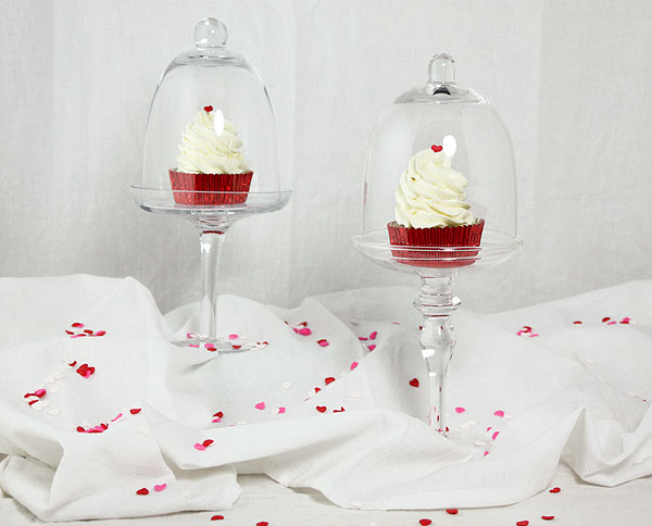 A photo of two Strawberry Cupcakes displayed in two glass cupcake stands with cloches on white cloth base with pink, white and red heart sprinkles.