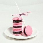 A photo of a stack of French Macarons filled with Raspberry Ganache displayed on a white dessert dish with French Macaron Cookie Pop leaning up against the stack.