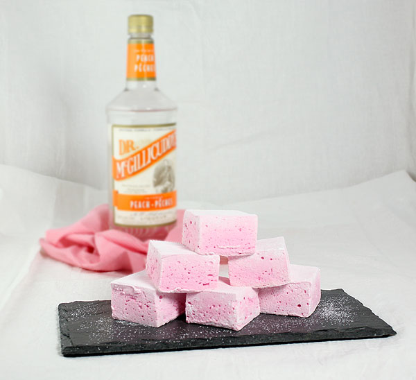 A photo of Boozy Strawberry Marshmallows stacked on a slate board with a bottle on Peach Schnapps set in behind on pink and white striped napkin.