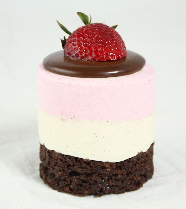 A close up photo of a Neapolitan Mousse Cake garnished with Dark Chocolate Ganache and fresh strawberry.