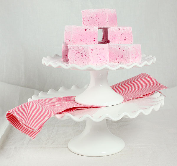 A photo of Strawberry Marshmallows stacked on small white cake stand that is stacked on a larger white cake stand with a pink and white striped napkin.
