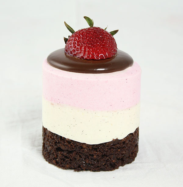 Close up photo of a Neapolitan Mousse Cake topped with a pool of dark chocolate ganache and fresh strawberry.
