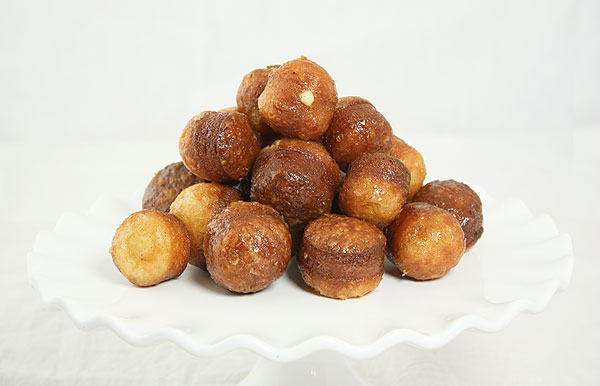 Close up photo of HOney Dipped and Vanilla Bean Cream filled Croissant Doughnut Holes stacked high on white cake stand.