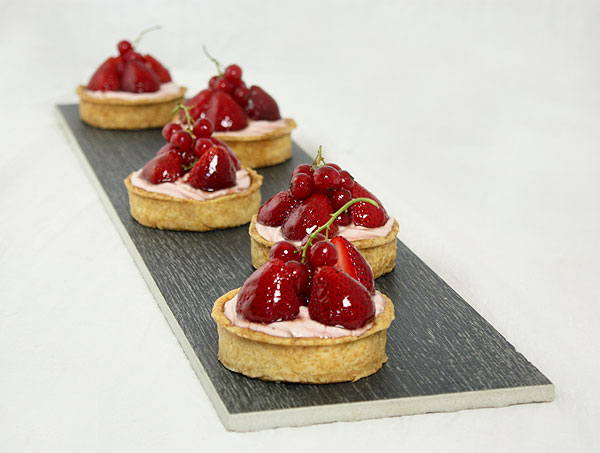 Photo of Cheesecake Tarts with Fresh Strawberries displayed in a row, one behind the other on rectangle shaped black ceramic plank.