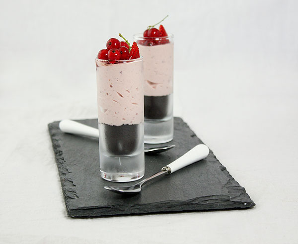 Photo of two Strawberry Cheesecake Verrines garnished with fresh strawberries and red currants displayed in shot glasses with dessert spoons on slate board.