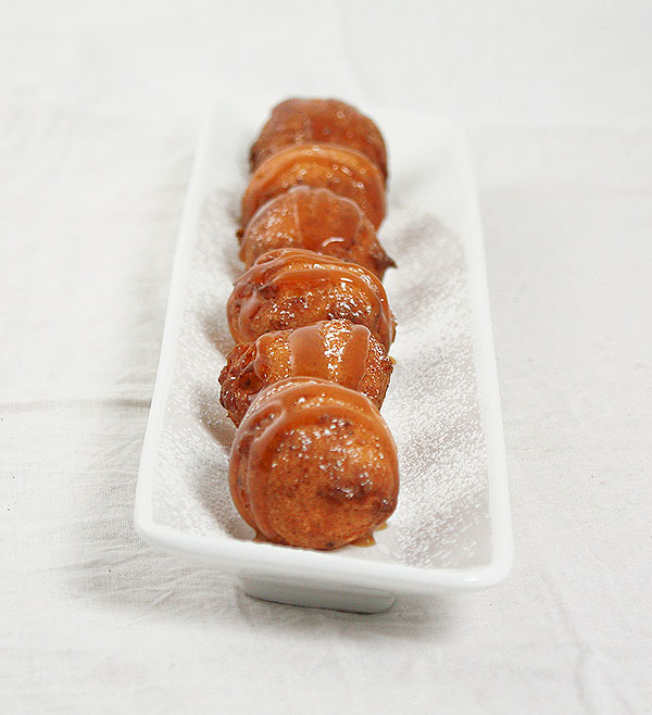 Photo of Sweet Ricotta Fritters dusted with confectioners' sugar and drizzled with butter-rum caramel sauce displayed on white rectangle-shaped serving dish displayed in a row, one behind the other.